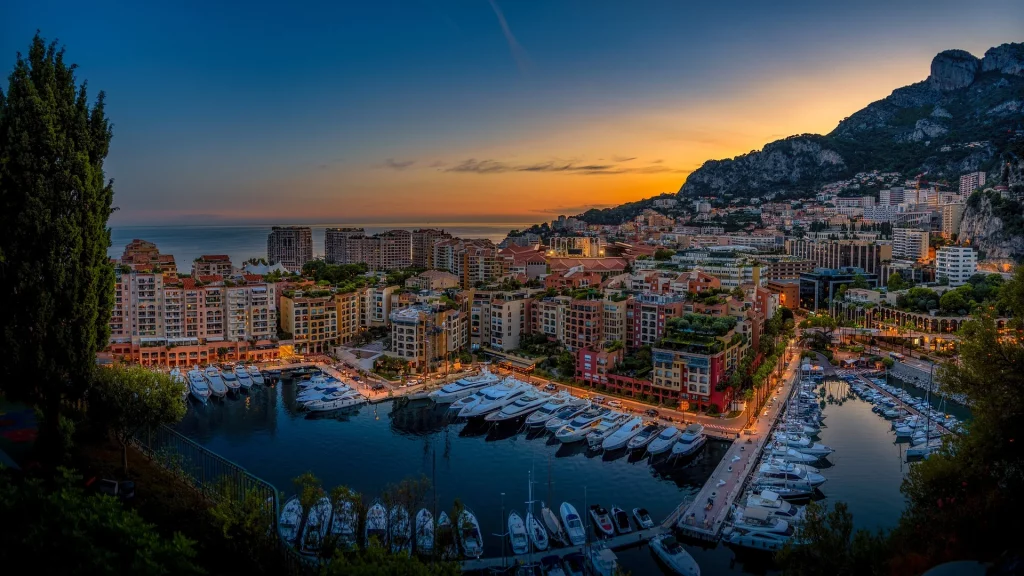 Vue panoramique du port de Monaco avec ses yachts luxueux, entouré de bâtiments élégants et des montagnes en arrière-plan.