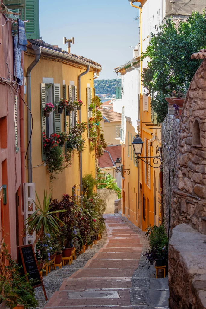 Vue panoramique de Menton, avec une route sinueuse bordée de végétation et un panneau indiquant l'entrée de la ville.