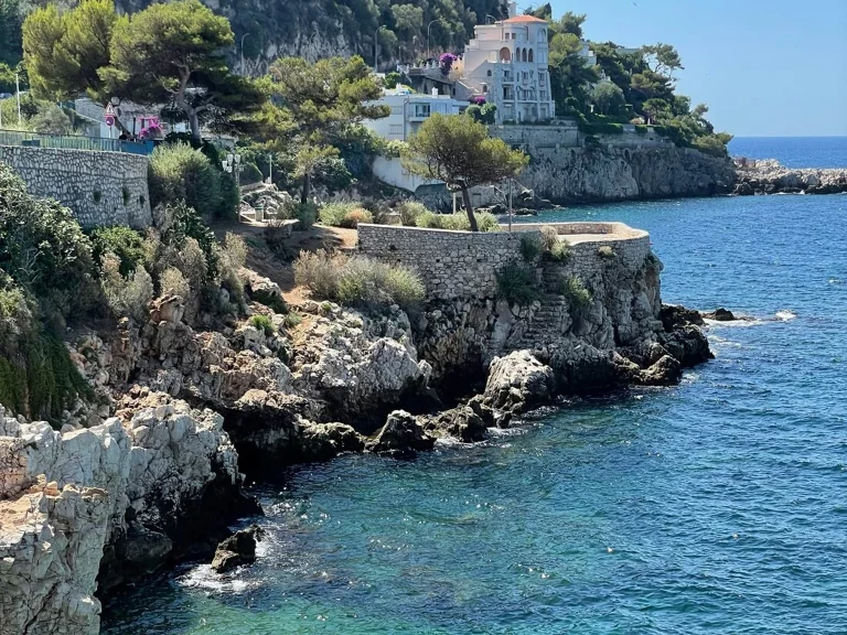 Vue panoramique de Nice, avec sa promenade des Anglais, ses plages bordées de galets et la mer Méditerranée scintillante sous un ciel bleu clair.